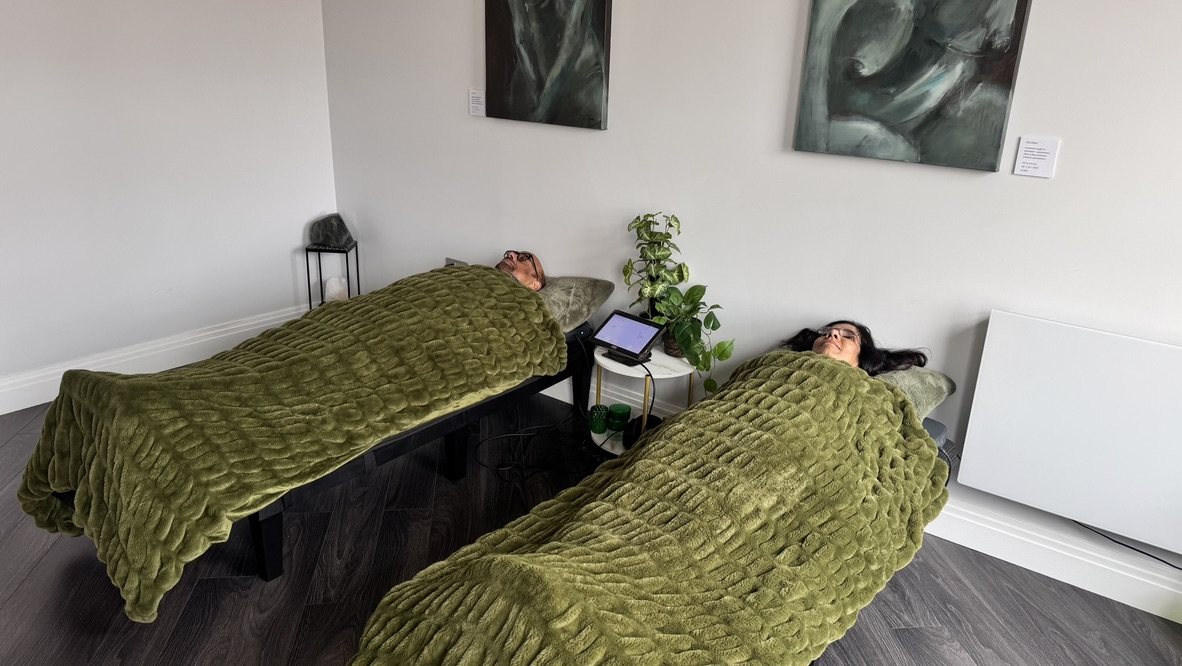 Two people resting side by side on the PEMF mats under green blankets in the quiet room